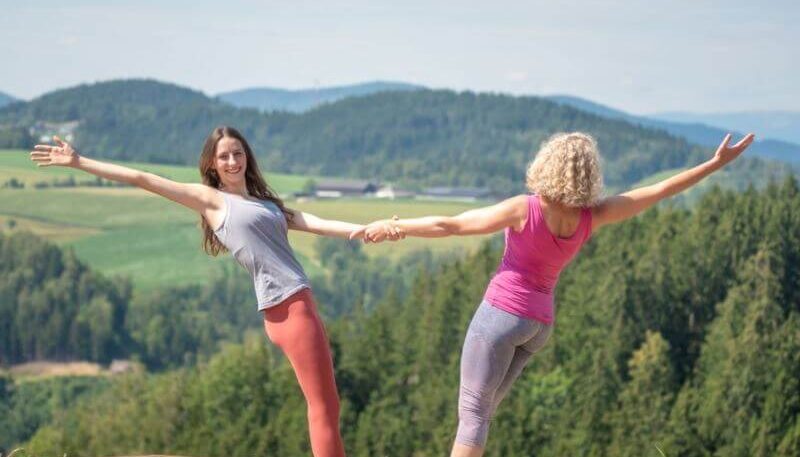 zwei Frauen greifen sich an den Armen und tanzen, im Hintergrund Wald und Wiesen