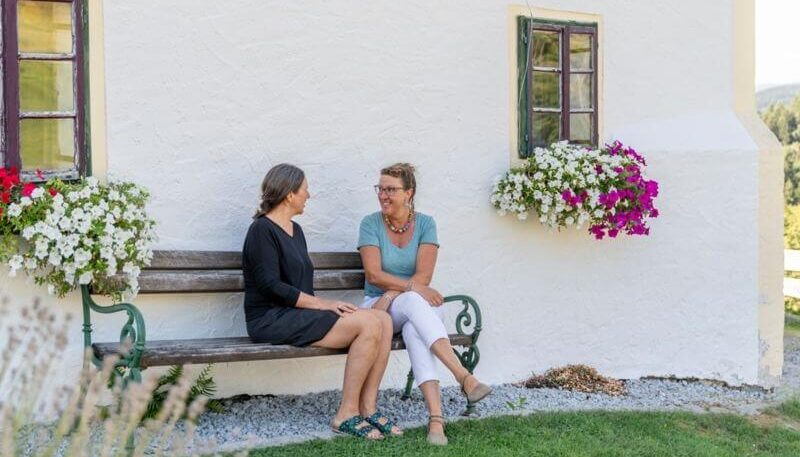 zwei Frauen sitzen auf eine Gartenbank an der Hausmauer und reden