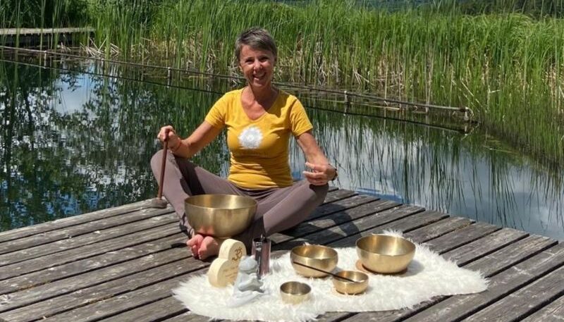 Frau meditiert mit Klangschalen am Wasser.
